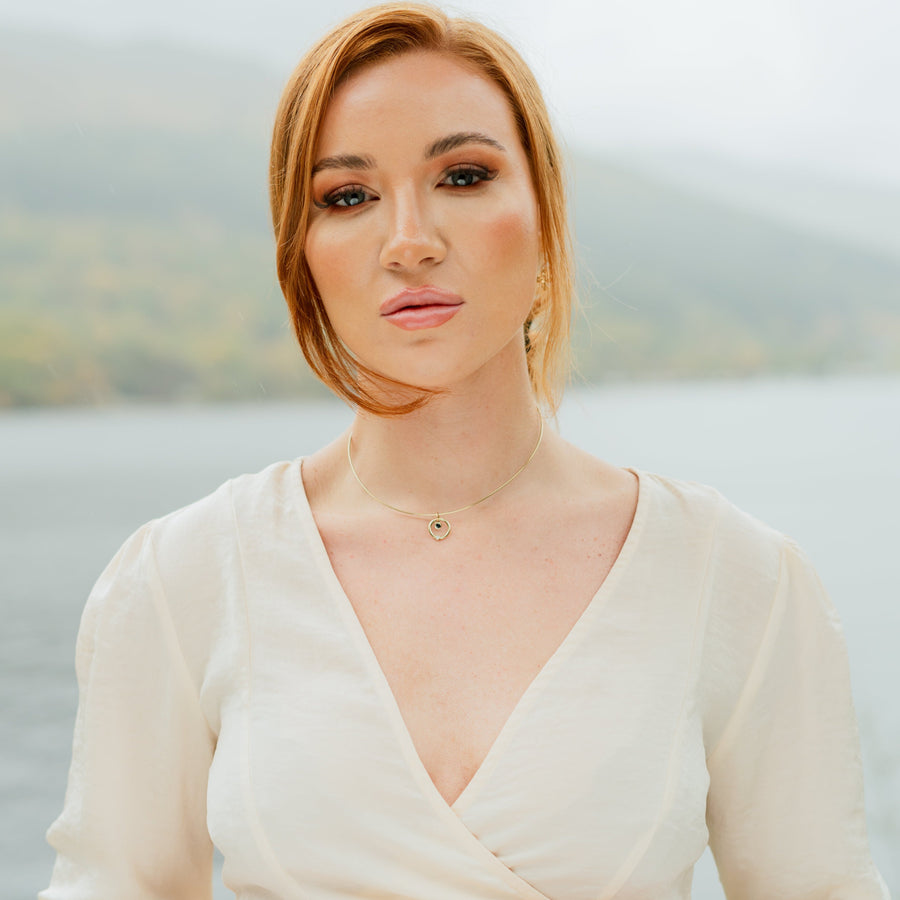 Pretty girl standing in front of a Scottish Loch. She's staring directly into the camera and wearing a white dress and a gold, diamond and sapphire solid necklace.