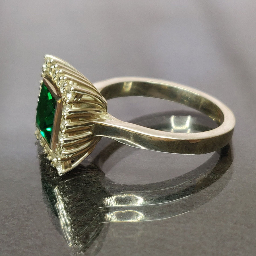 Profile view of a princess cut emerald and white diamond halo cluster ring on a reflective surface.