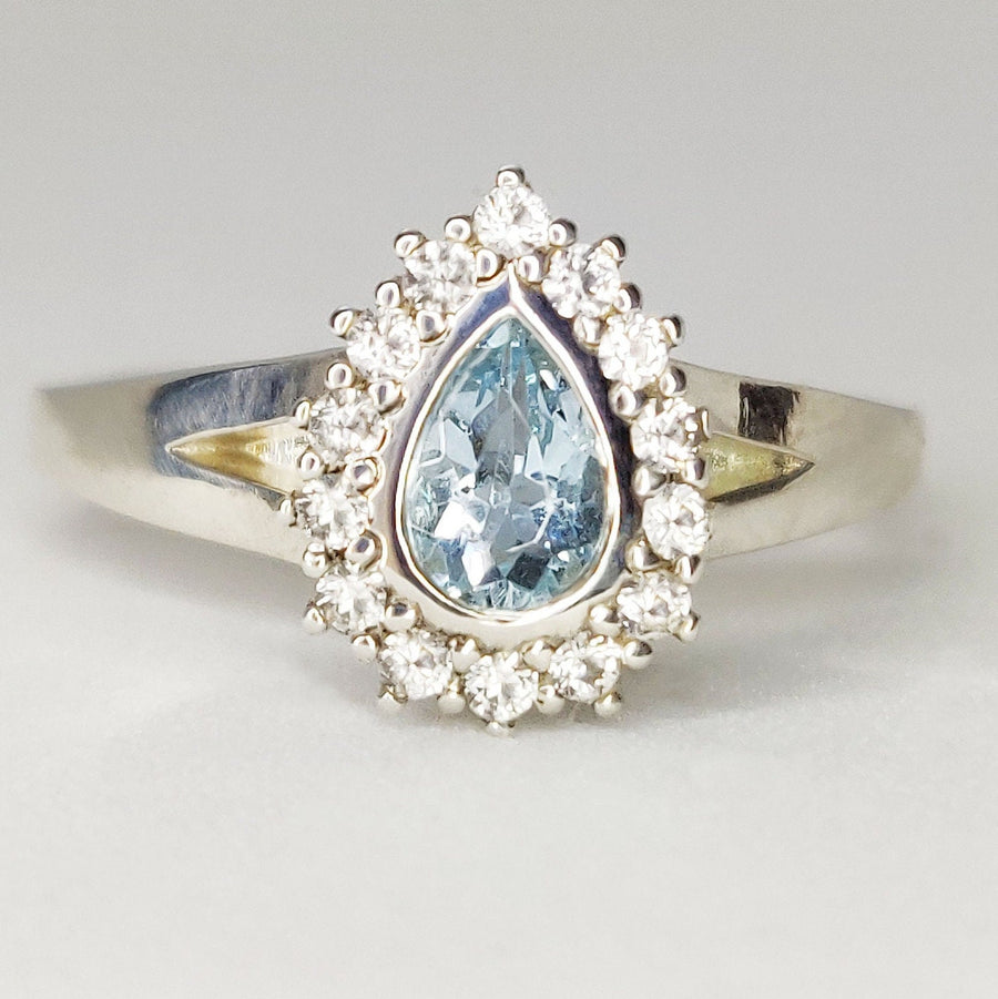Pear cut aquamarine and white sapphire halo cluster ring on a white surface.