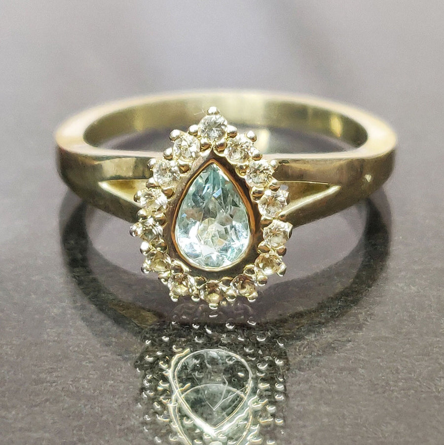 Pear cut aquamarine and white sapphire halo cluster ring on a reflective surface.