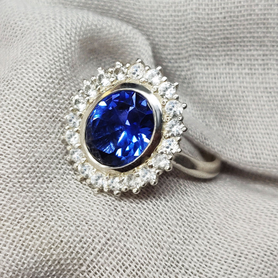 Front view of a bezel set blue and white sapphire halo cluster ring on a canvas background.