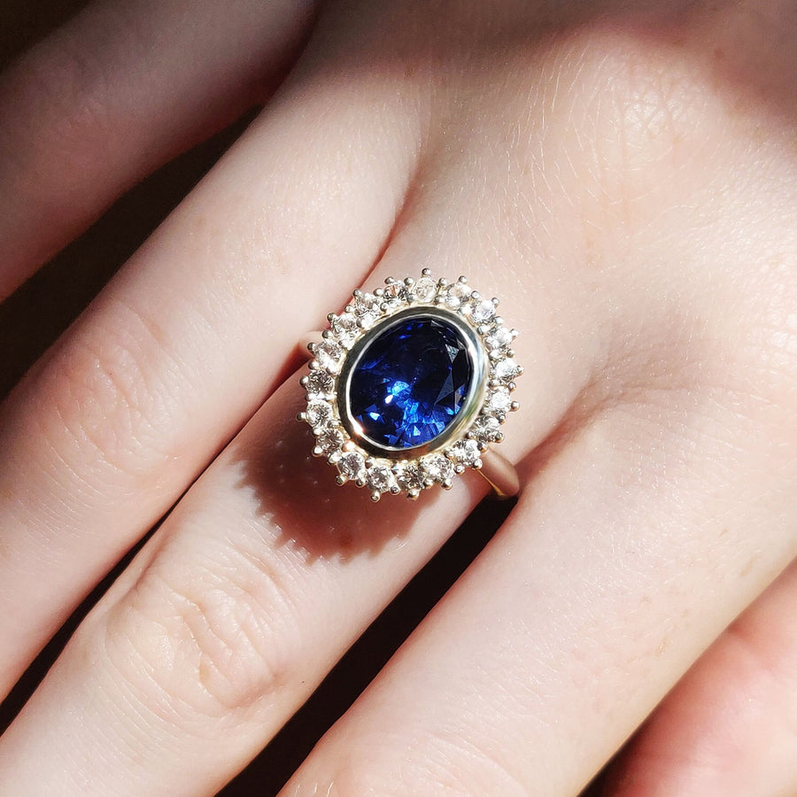 Bezel set blue and white sapphire halo cluster ring being worn on a finger.