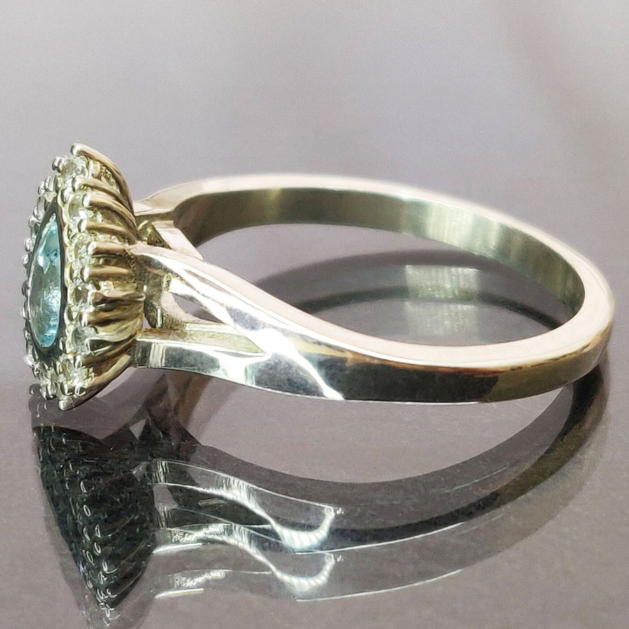 Side view of a pear cut aquamarine and white sapphire halo cluster ring on a reflective surface.