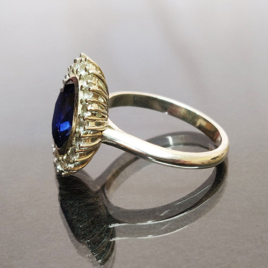 Side view of a bezel set blue and white sapphire halo cluster ring on a reflective surface.