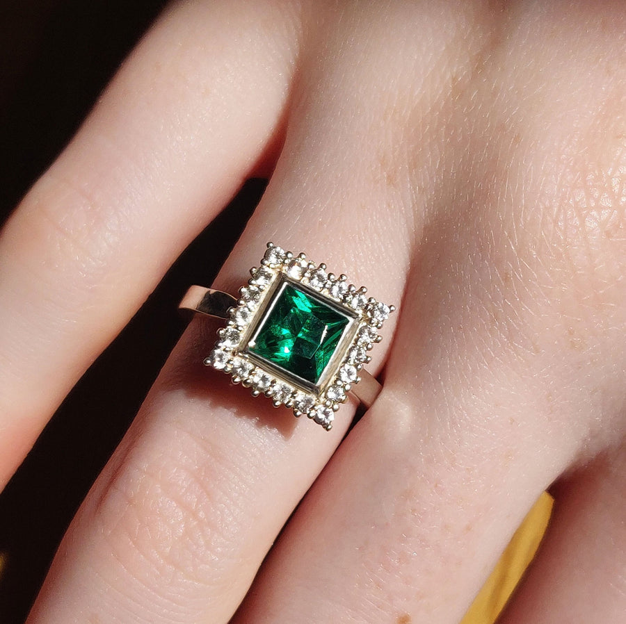 princess cut emerald and white diamond halo cluster ring being worn on a finger.