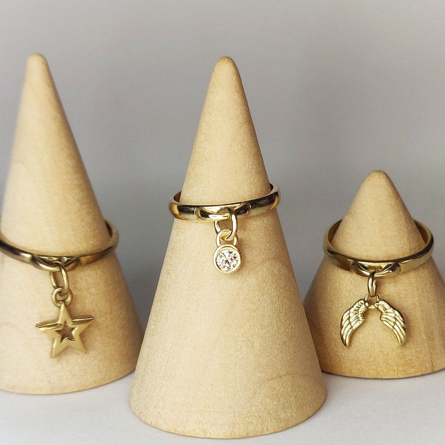 3 Charm rings with diamond, angel wings and star charms, all on wooden displays against a white background.