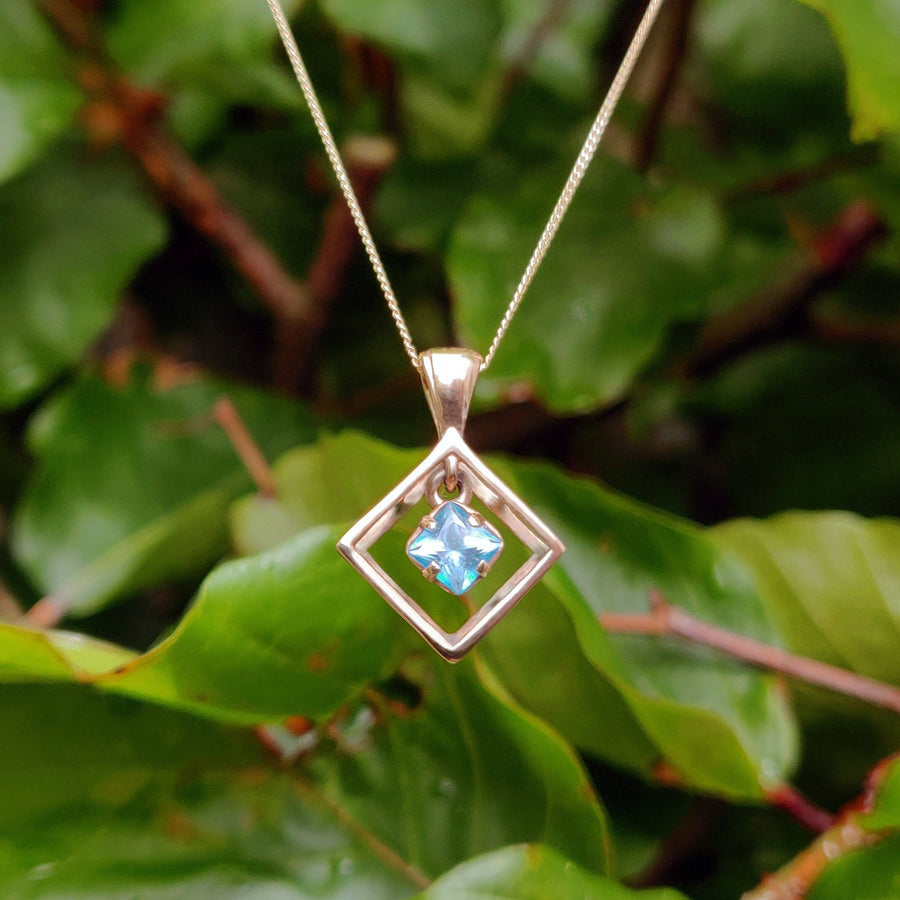 Blue princess cut sapphire in a gold necklace, hanging against a nature background.